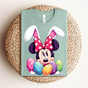 May include: A pale green t-shirt with a Minnie Mouse cartoon design, complete with bunny ears and a red polka dot bow. The design includes colourful Easter eggs. The shirt is on a woven, circular placemat.