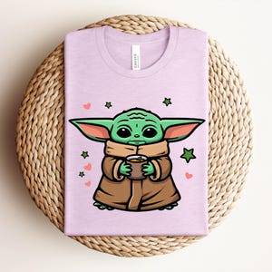 May include: A light purple t-shirt featuring a cartoon character with large green ears, holding a cup. The character is wearing a brown coat and a tan scarf. Green stars and pink hearts are scattered around the character.