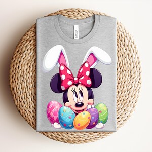 May include: A grey t-shirt featuring a cartoon Minnie Mouse design with bunny ears and a red polka dot bow. The design includes colourful Easter eggs. The shirt is folded on a woven surface.