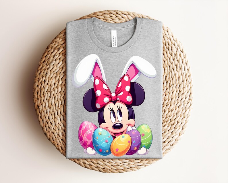 May include: A grey t-shirt featuring a cartoon Minnie Mouse design with bunny ears and a red polka dot bow. The design includes colourful Easter eggs. The shirt is folded on a woven surface.