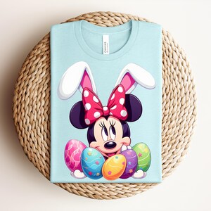 May include: A pale blue t-shirt featuring Minnie Mouse with bunny ears and a red polka dot bow. The graphic includes colourful Easter eggs. The shirt is folded on a woven, circular surface.