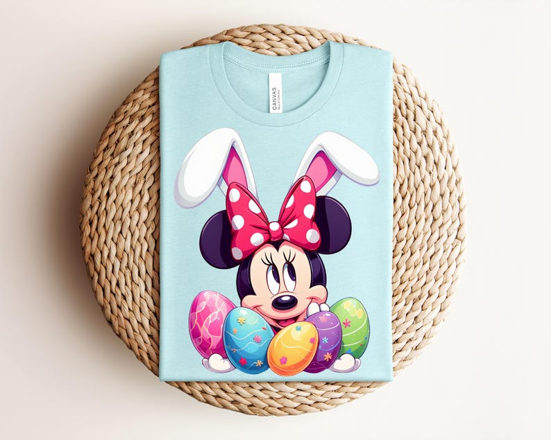 May include: A pale blue t-shirt featuring Minnie Mouse with bunny ears and a red polka dot bow. The graphic includes colourful Easter eggs. The shirt is folded on a woven, circular surface.