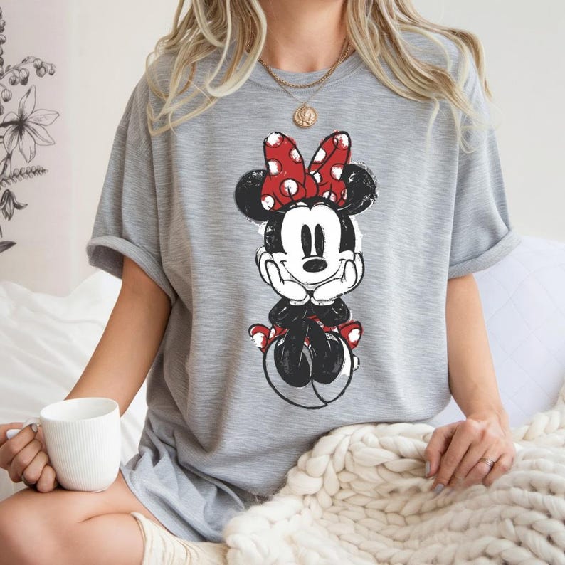 May include: A grey t-shirt with a distressed graphic of Minnie Mouse. Minnie is shown with a red polka-dot bow, black ears, and a black and white outfit. The t-shirt has short sleeves and a relaxed fit.
