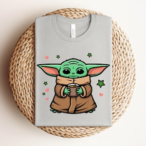 May include: A light gray t-shirt featuring a cartoon illustration of a green alien character holding a cup. The character is wearing a brown coat and surrounded by green stars and pink hearts.