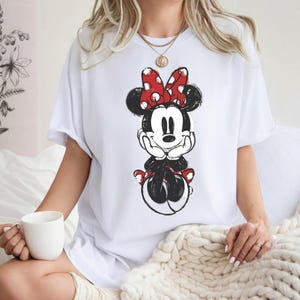 May include: White t-shirt with a distressed Minnie Mouse graphic. The design features Minnie in black, red, and white, with a red polka-dot bow. The shirt has short sleeves and a relaxed fit, suitable for casual wear.