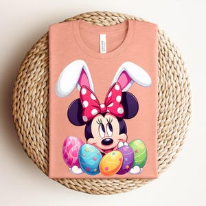 May include: Peach-coloured t-shirt featuring a Minnie Mouse graphic with bunny ears and a polka-dot bow. The design includes colourful Easter eggs. The shirt is folded on a woven, circular surface.