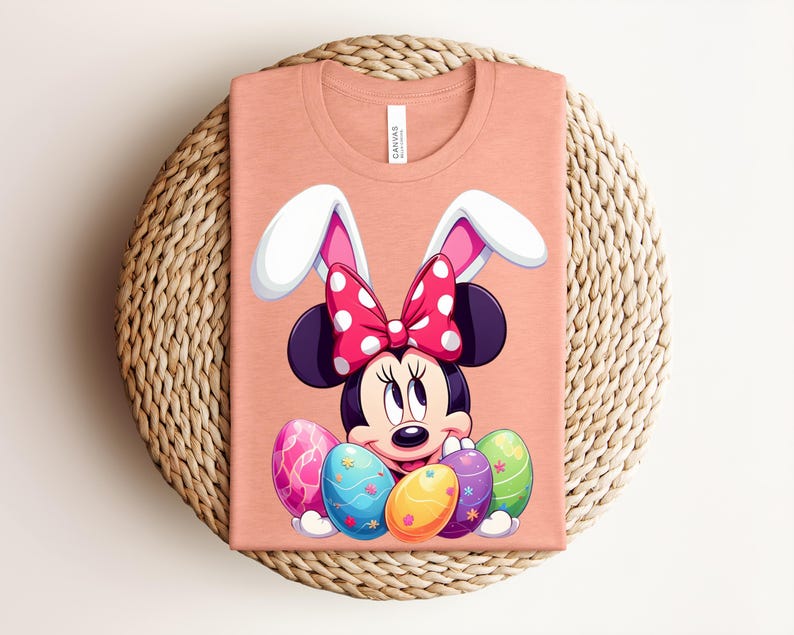 May include: Peach-coloured t-shirt featuring a Minnie Mouse graphic with bunny ears and a polka-dot bow. The design includes colourful Easter eggs. The shirt is folded on a woven, circular surface.