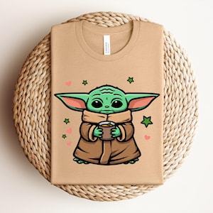 May include: A tan t-shirt featuring a cartoon illustration of a green alien character holding a cup. The character is wearing a brown cloak, with green ears and large black eyes. Pink hearts and green stars surround the character.