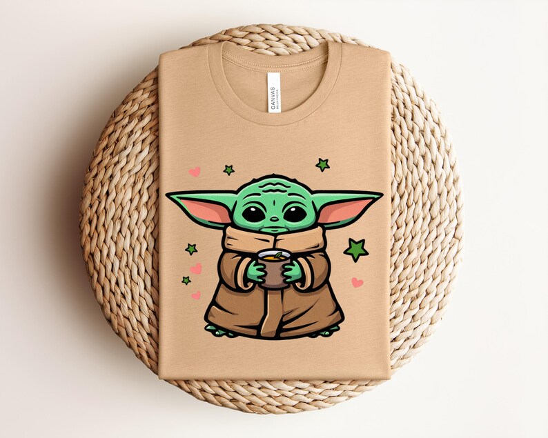 May include: A tan t-shirt featuring a cartoon illustration of a green alien character holding a cup. The character is wearing a brown cloak, with green ears and large black eyes. Pink hearts and green stars surround the character.