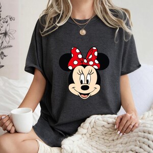 Op de afbeelding: Donkergrijs T-shirt met een Minnie Mouse-afbeelding. De afbeelding toont Minnie's gezicht met een rode strik met witte stippen. Het shirt heeft korte mouwen en een ronde hals.