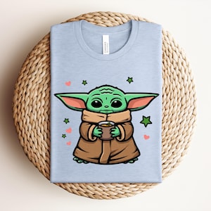 May include: Light blue t-shirt featuring a cartoon illustration of a green alien character holding a cup. The character is wearing a brown cloak and has large ears. The design includes green stars and pink hearts.
