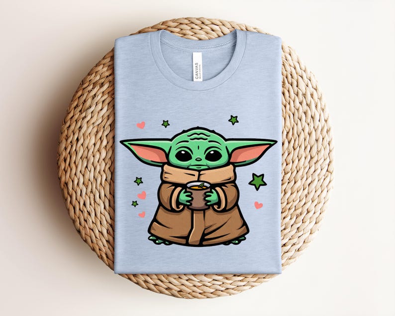 May include: Light blue t-shirt featuring a cartoon illustration of a green alien character holding a cup. The character is wearing a brown cloak and has large ears. The design includes green stars and pink hearts.