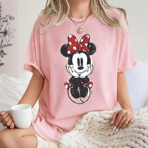 May include: A pale pink t-shirt with a Minnie Mouse graphic. The cartoon character is in black and white with a red polka dot bow. The top has short sleeves and a loose fit, ideal for everyday wear.