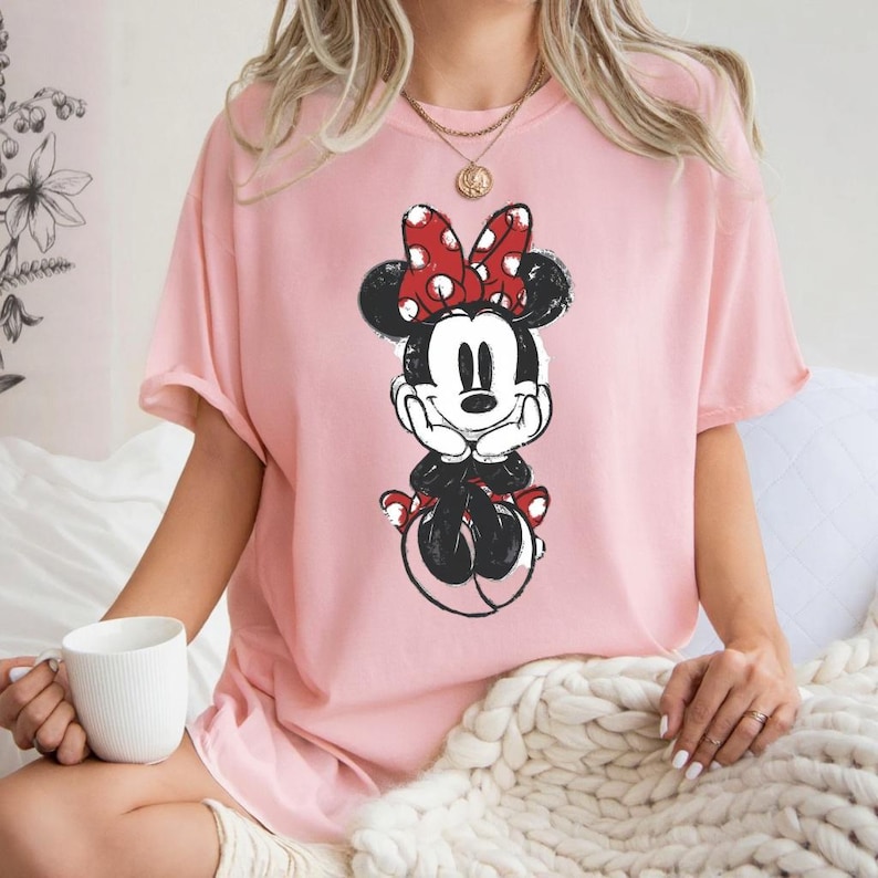 May include: A pale pink t-shirt with a Minnie Mouse graphic. The cartoon character is in black and white with a red polka dot bow. The top has short sleeves and a loose fit, ideal for everyday wear.