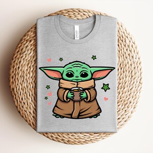 May include: A gray t-shirt featuring a cartoon character holding a cup. The character is green with large ears and is wearing a brown coat. Pink hearts and green stars are scattered around the character.