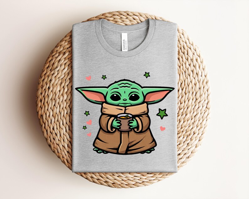 May include: A gray t-shirt featuring a cartoon character holding a cup. The character is green with large ears and is wearing a brown coat. Pink hearts and green stars are scattered around the character.