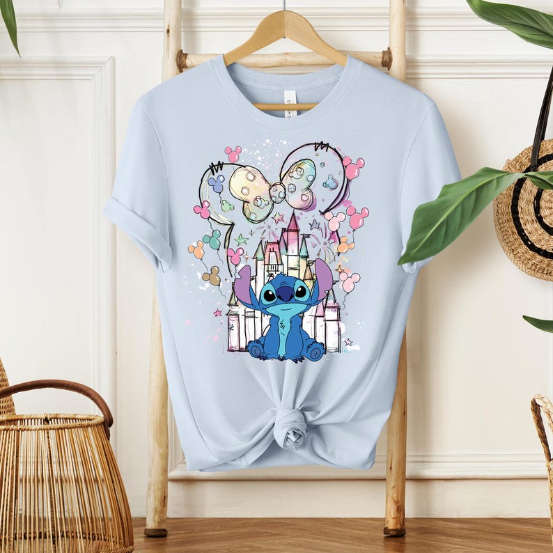 K&ouml;nnte beinhalten: Hellblaues T-Shirt mit einem farbenfrohen Aufdruck von Stitch aus Lilo & Stitch, einem Schloss und Mickey Mouse Ohren. Das Design verwendet Pastellfarben und Sterne. Das T-Shirt h&auml;ngt an einem Holzb&uuml;gel.