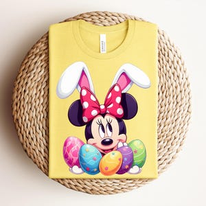 May include: Yellow t-shirt featuring Minnie Mouse as an Easter bunny, with white and pink bunny ears and a red polka dot bow. Minnie Mouse is surrounded by colourful Easter eggs. The shirt is folded on a woven, circular surface.