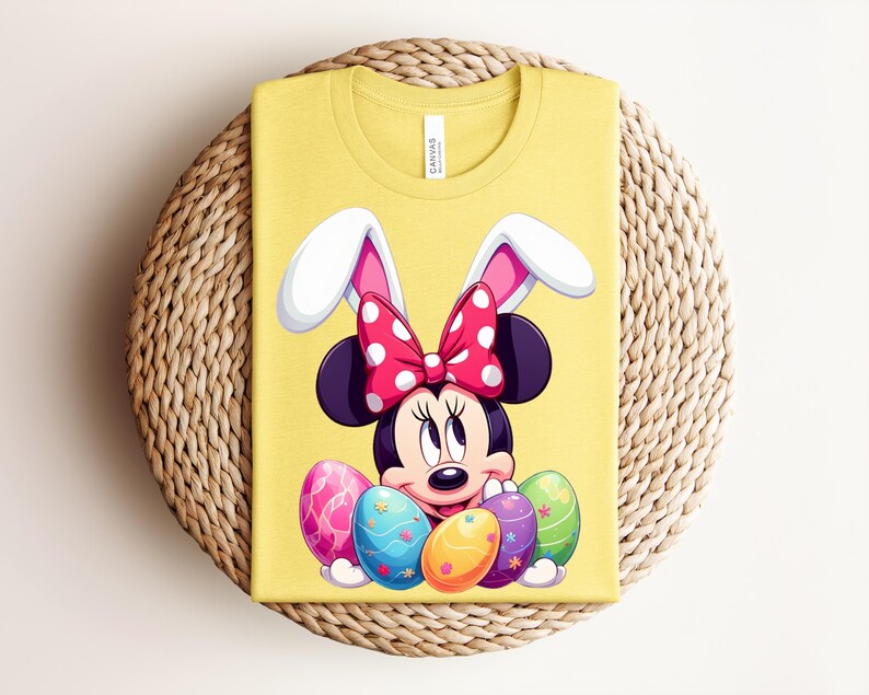 May include: Yellow t-shirt featuring Minnie Mouse as an Easter bunny, with white and pink bunny ears and a red polka dot bow. Minnie Mouse is surrounded by colourful Easter eggs. The shirt is folded on a woven, circular surface.