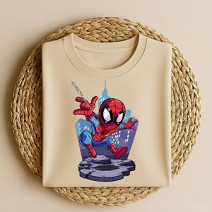 K&ouml;nnte beinhalten: Beiges Rundhals-T-Shirt mit einem Cartoon-Spider-Man-Motiv. Die Illustration zeigt Spider-Man in seinem roten und blauen Anzug, der vor einer Skyline schwingt. Das Design befindet sich auf einer steinartigen Plattform.