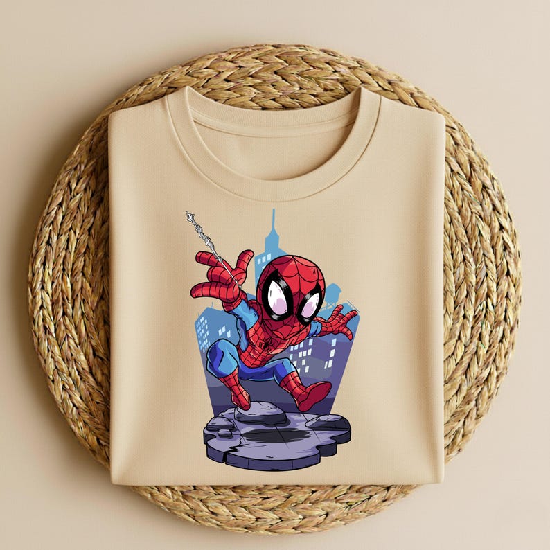 K&ouml;nnte beinhalten: Beiges Rundhals-T-Shirt mit einem Cartoon-Spider-Man-Motiv. Die Illustration zeigt Spider-Man in seinem roten und blauen Anzug, der vor einer Skyline schwingt. Das Design befindet sich auf einer steinartigen Plattform.