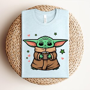 May include: Light blue t-shirt featuring a cartoon character holding a cup. The character has green skin, large ears, and is wearing a brown cloak. The design includes green stars and pink hearts. The shirt is folded on a woven surface.