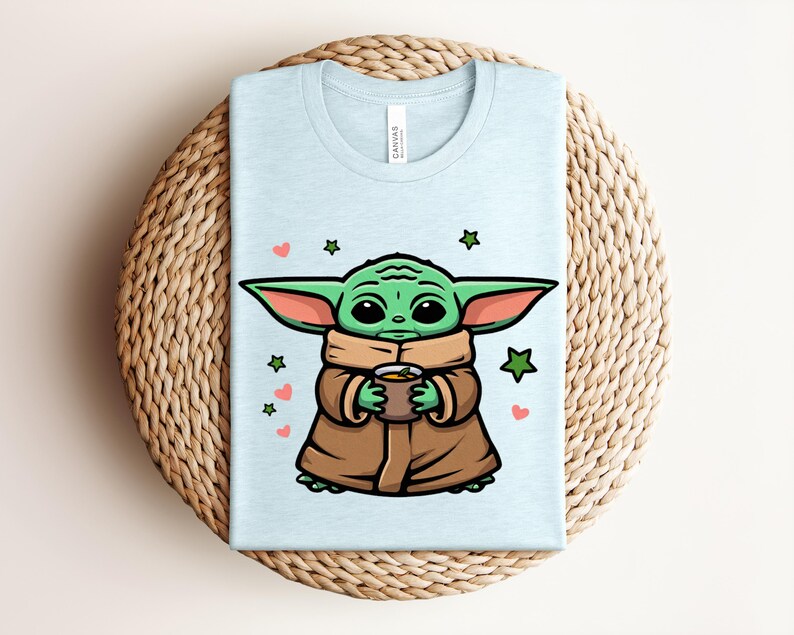 May include: Light blue t-shirt featuring a cartoon character holding a cup. The character has green skin, large ears, and is wearing a brown cloak. The design includes green stars and pink hearts. The shirt is folded on a woven surface.
