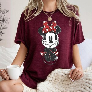 May include: Burgundy t-shirt with a Minnie Mouse graphic. The cartoon character is in black, white, and red, with a large polka-dot bow. The shirt has short sleeves and a relaxed fit, suitable for casual wear.