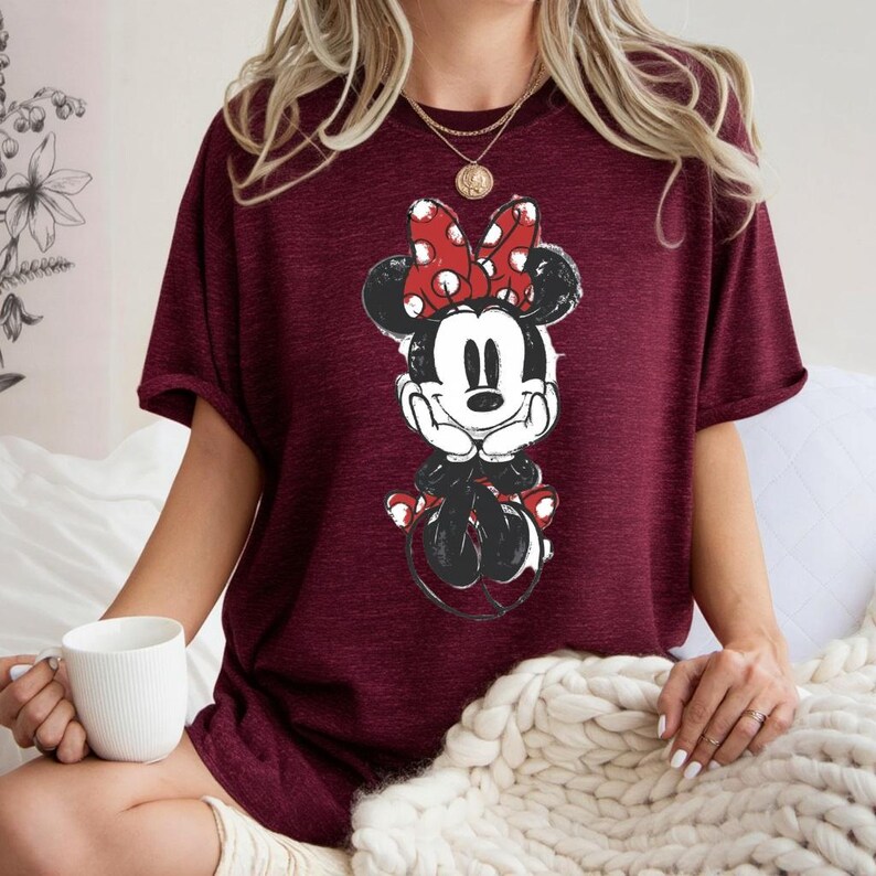May include: Burgundy t-shirt with a Minnie Mouse graphic. The cartoon character is in black, white, and red, with a large polka-dot bow. The shirt has short sleeves and a relaxed fit, suitable for casual wear.