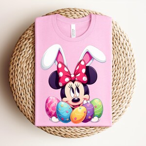 May include: Pink t-shirt featuring Minnie Mouse as an Easter bunny. Minnie wears a red polka dot bow and white bunny ears. Colourful Easter eggs are in front of her. The shirt is on a woven, circular surface.