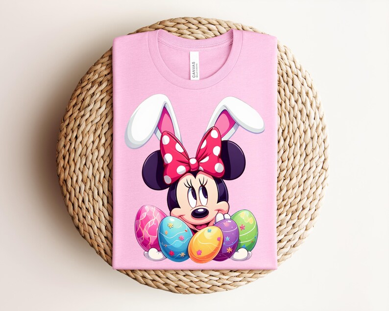 May include: Pink t-shirt featuring Minnie Mouse as an Easter bunny. Minnie wears a red polka dot bow and white bunny ears. Colourful Easter eggs are in front of her. The shirt is on a woven, circular surface.