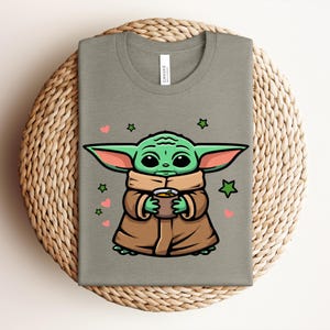May include: A sage green t-shirt featuring a cartoon character holding a cup. The character has large green ears, big eyes, and is wearing a brown coat. Pink hearts and green stars surround the character. The shirt is folded on a woven mat.