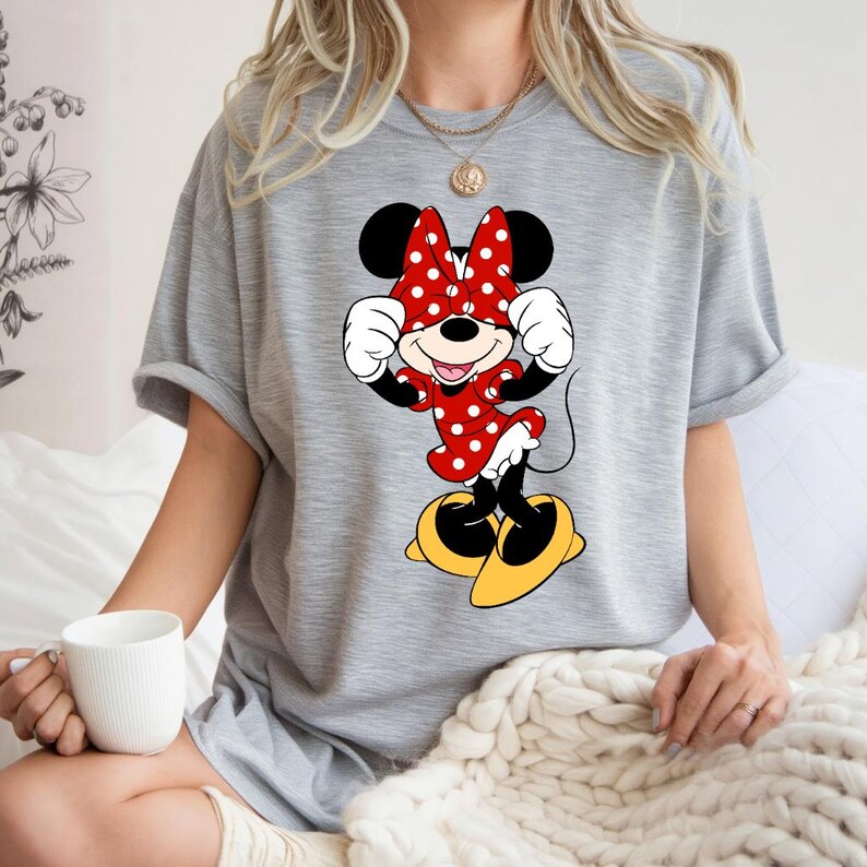 K&ouml;nnte beinhalten: Ein graues T-Shirt mit einem Minnie Mouse-Motiv. Minnie Mouse ist in einem roten Polka-Dot-Kleid, passender Schleife und gelben Schuhen dargestellt. Die Zeichentrickfigur l&auml;chelt mit erhobenen Armen. Das T-Shirt ist ein l&auml;ssiger Stil.