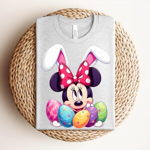 May include: A light grey t-shirt featuring a cartoon Minnie Mouse design with bunny ears and a red polka dot bow. The design includes colourful Easter eggs. The shirt is folded on a woven surface.