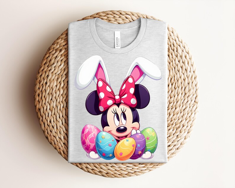 May include: A light grey t-shirt featuring a cartoon Minnie Mouse design with bunny ears and a red polka dot bow. The design includes colourful Easter eggs. The shirt is folded on a woven surface.