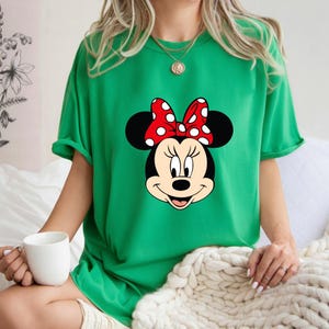 Op de afbeelding: Een groen t-shirt met een Minnie Mouse-afbeelding. De afbeelding toont Minnie's gezicht met een rode strik met witte stippen. Het t-shirt is effen groen van kleur en heeft korte mouwen.