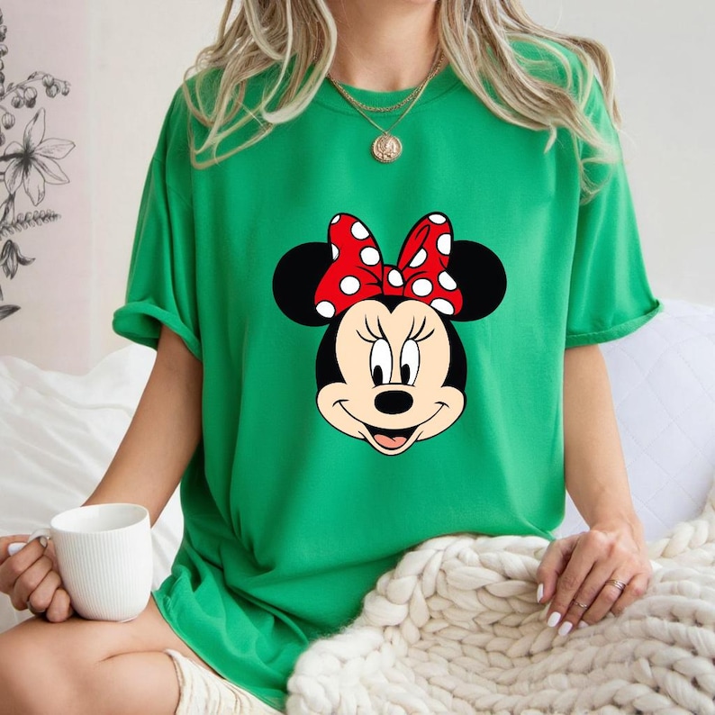 Op de afbeelding: Een groen t-shirt met een Minnie Mouse-afbeelding. De afbeelding toont Minnie's gezicht met een rode strik met witte stippen. Het t-shirt is effen groen van kleur en heeft korte mouwen.