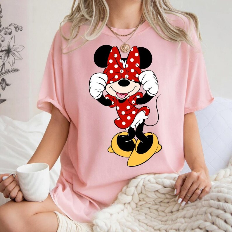 K&ouml;nnte beinhalten: Hellrosa T-Shirt mit Minnie Mouse-Grafik. Minnie ist in einem roten Kleid mit wei&szlig;en Punkten, einer passenden Schleife und gelben Schuhen dargestellt. Die Zeichentrickfigur l&auml;chelt und bedeckt ihre Augen mit ihren H&auml;nden.