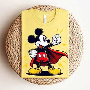 May include: Yellow t-shirt featuring a cartoon illustration of Mickey Mouse in a superhero pose. Mickey is wearing a red cape, shorts, and yellow shoes. The shirt is folded on a woven, circular surface.
