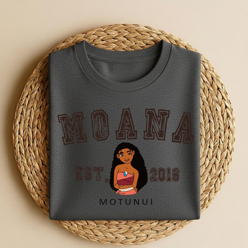 Puede incluir: Camiseta gris oscuro de cuello redondo con la palabra "MOANA" en letras marrones en bloque sobre una ilustraci&oacute;n de dibujos animados de una mujer. El texto "EST. 2016" y "MOTUNUI" tambi&eacute;n est&aacute;n impresos en la camiseta.
