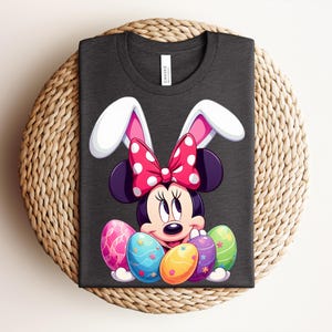 May include: Dark grey t-shirt featuring Minnie Mouse with bunny ears and a polka dot bow. Minnie is surrounded by colourful Easter eggs. The shirt is folded on a woven, circular surface.