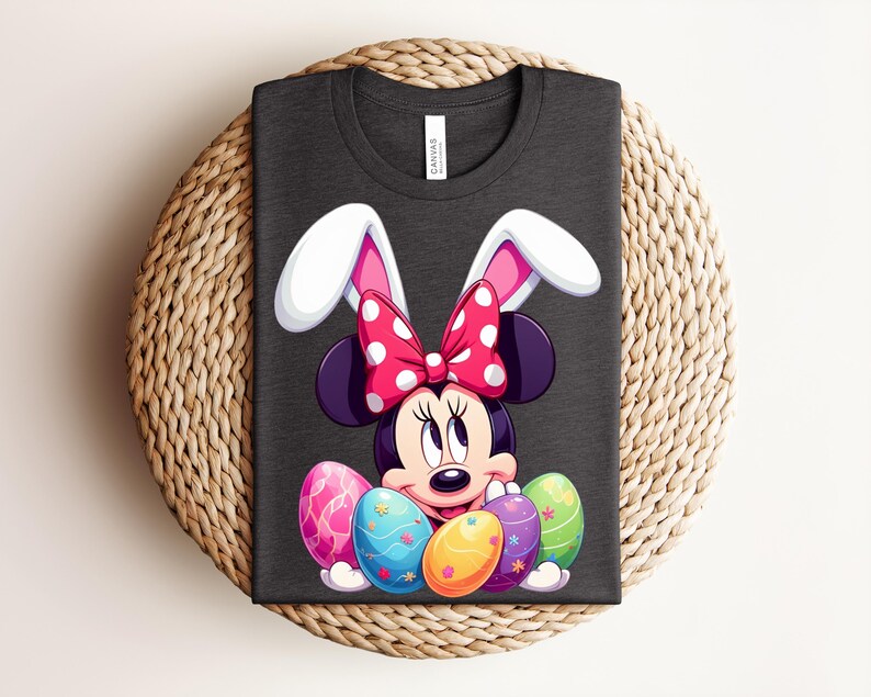 May include: Dark grey t-shirt featuring Minnie Mouse with bunny ears and a polka dot bow. Minnie is surrounded by colourful Easter eggs. The shirt is folded on a woven, circular surface.