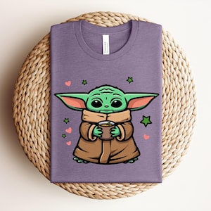 May include: A purple t-shirt featuring a cartoon character holding a cup. The character has green skin, large ears, and is wearing a brown cloak. Pink hearts and green stars are scattered around the character.