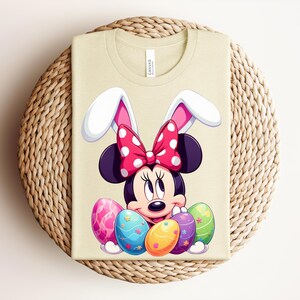 May include: A light beige t-shirt featuring a Minnie Mouse design with bunny ears and a polka dot bow. The design includes colourful Easter eggs. The shirt is folded on a woven, circular surface.