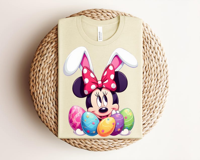 May include: A light beige t-shirt featuring a Minnie Mouse design with bunny ears and a polka dot bow. The design includes colourful Easter eggs. The shirt is folded on a woven, circular surface.