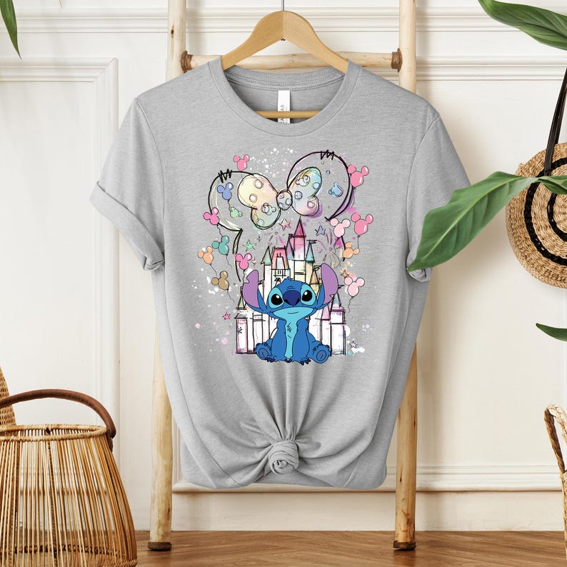 K&ouml;nnte beinhalten: Hellgraues T-Shirt mit einem Cartoon-Stitch-Charakter vor einem Schloss. Das Design umfasst eine Schleife und Mickey Mouse-f&ouml;rmige Ballons in Pastellfarben. Das T-Shirt h&auml;ngt an einem Holzb&uuml;gel.