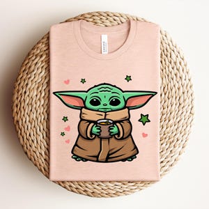 May include: A light pink t-shirt featuring a cartoon character with large green ears, holding a cup. The character is wearing a brown coat and a tan scarf. Green stars and pink hearts are scattered around the character.