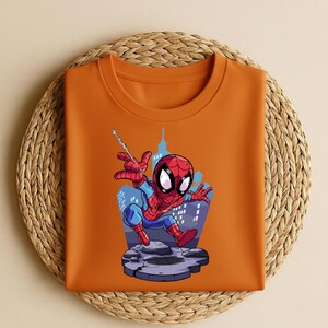 K&ouml;nnte beinhalten: Ein orangefarbenes T-Shirt mit Rundhalsausschnitt und einem Spider-Man-Cartoon-Motiv. Der Superheld ist in seinem rot-blauen Anzug dargestellt, mit einem Stadtlandschaftshintergrund. Das T-Shirt ist auf einem gewebten, kreisf&ouml;rmigen Platzset gefaltet.
