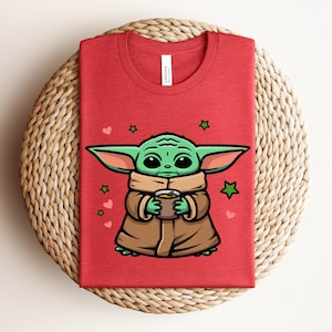 May include: Red t-shirt featuring a cartoon illustration of a green alien character holding a cup. The character is wearing a brown robe and scarf, with green stars and pink hearts around it. The shirt is folded on a woven surface.