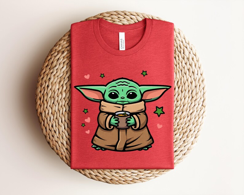 May include: Red t-shirt featuring a cartoon illustration of a green alien character holding a cup. The character is wearing a brown robe and scarf, with green stars and pink hearts around it. The shirt is folded on a woven surface.